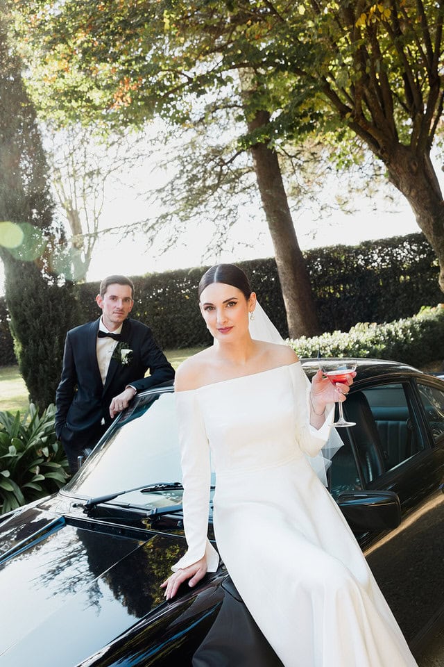 Fashion‑inspired car portrait at Les Jardins de Campagne – Maxime Bernadin. Bride sits on a vintage black sports car holding a cocktail while the groom leans on the hood at Les Jardins de Campagne, captured by Maxime Bernadin.