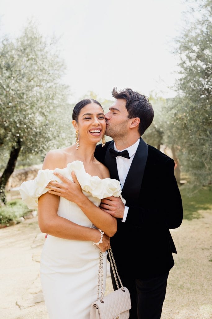 Olive grove laughter at Château de Berne – Maxime Bernadin. Groom kisses bride’s cheek as she laughs in an olive grove at Château de Berne, captured by Maxime Bernadin.