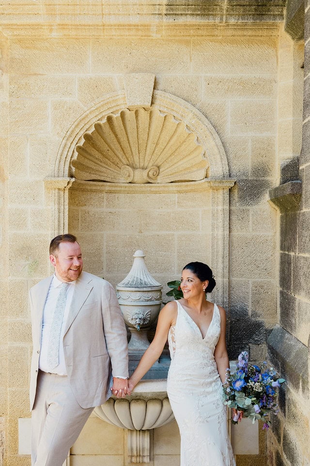 Stone niche portrait at Le Vieux Castillon – Maxime Bernadin Bride in lace dress with blue bouquet and groom in beige suit stand by a stone niche at Le Vieux Castillon, captured by Maxime Bernadin.