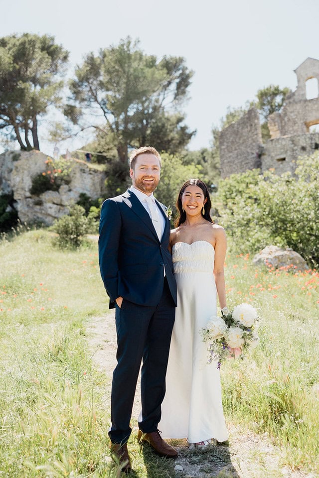 Wildflower meadow romance at Mas de la Rose – Maxime Bernadin. Bride in strapless gown and groom in navy suit stand in a wildflower meadow with stone ruins at Mas de la Rose, captured by Maxime Bernadin