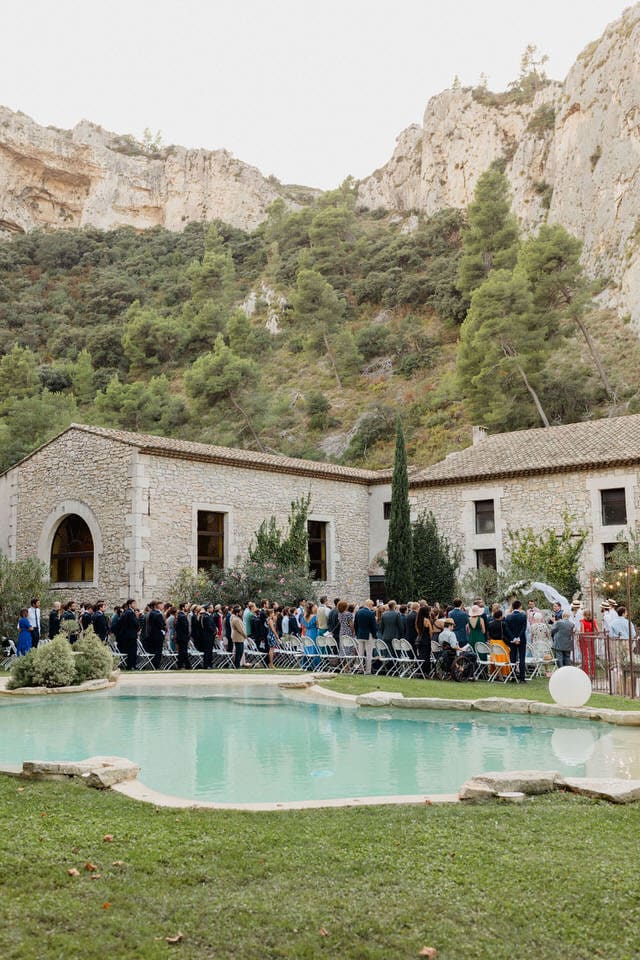 Poolside ceremony at Domaine du Rocher – Maxime Bernadin Guests seated at an outdoor ceremony beside a turquoise pool and stone estate beneath limestone cliffs at Domaine du Rocher, captured by Maxime Bernadin