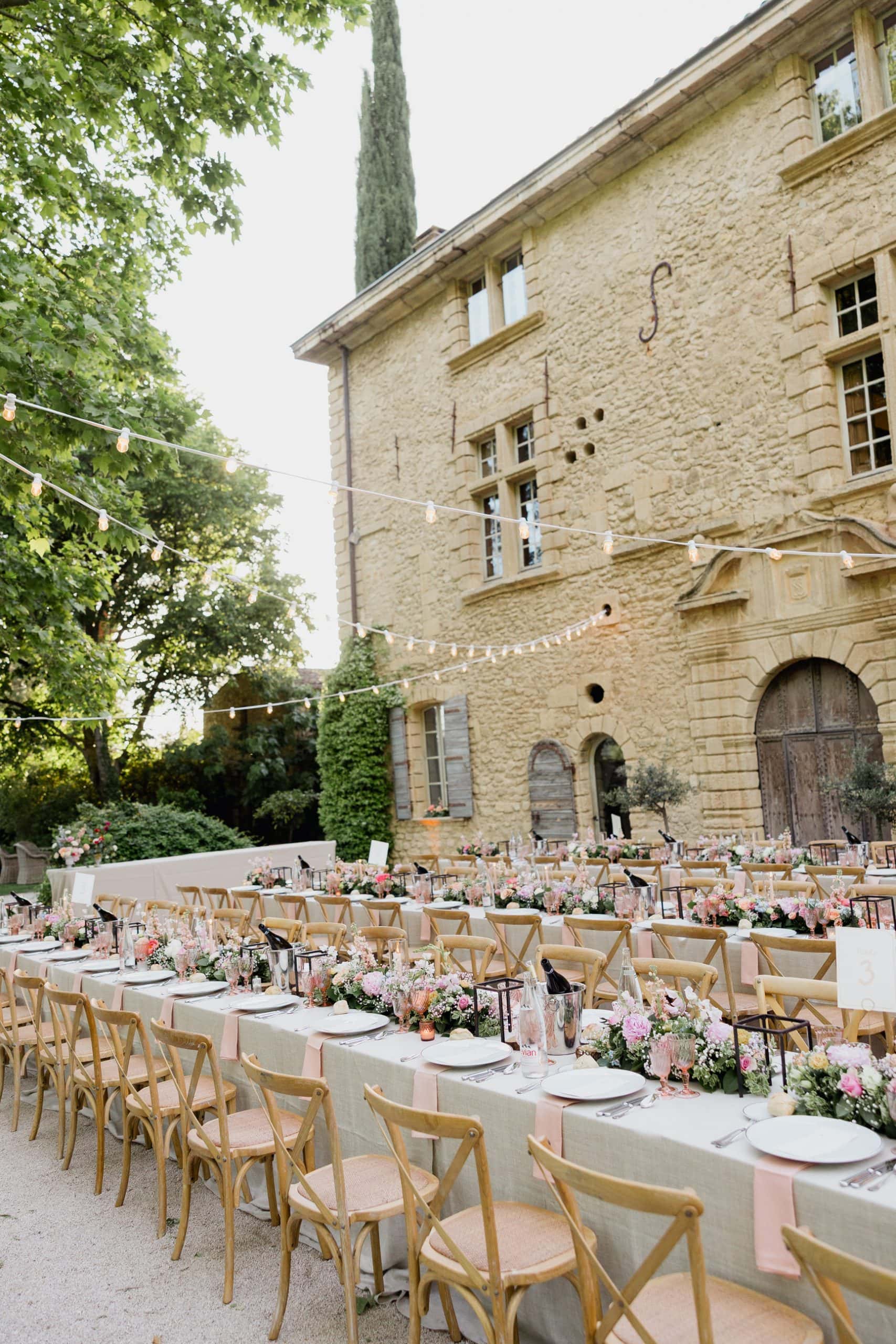 Outdoor banquet at Château de Sannes – Maxime Bernadin. Long banquet tables adorned with pastel flowers and string lights outside a stone manor at Château de Sannes, photographed by Maxime Bernadin.