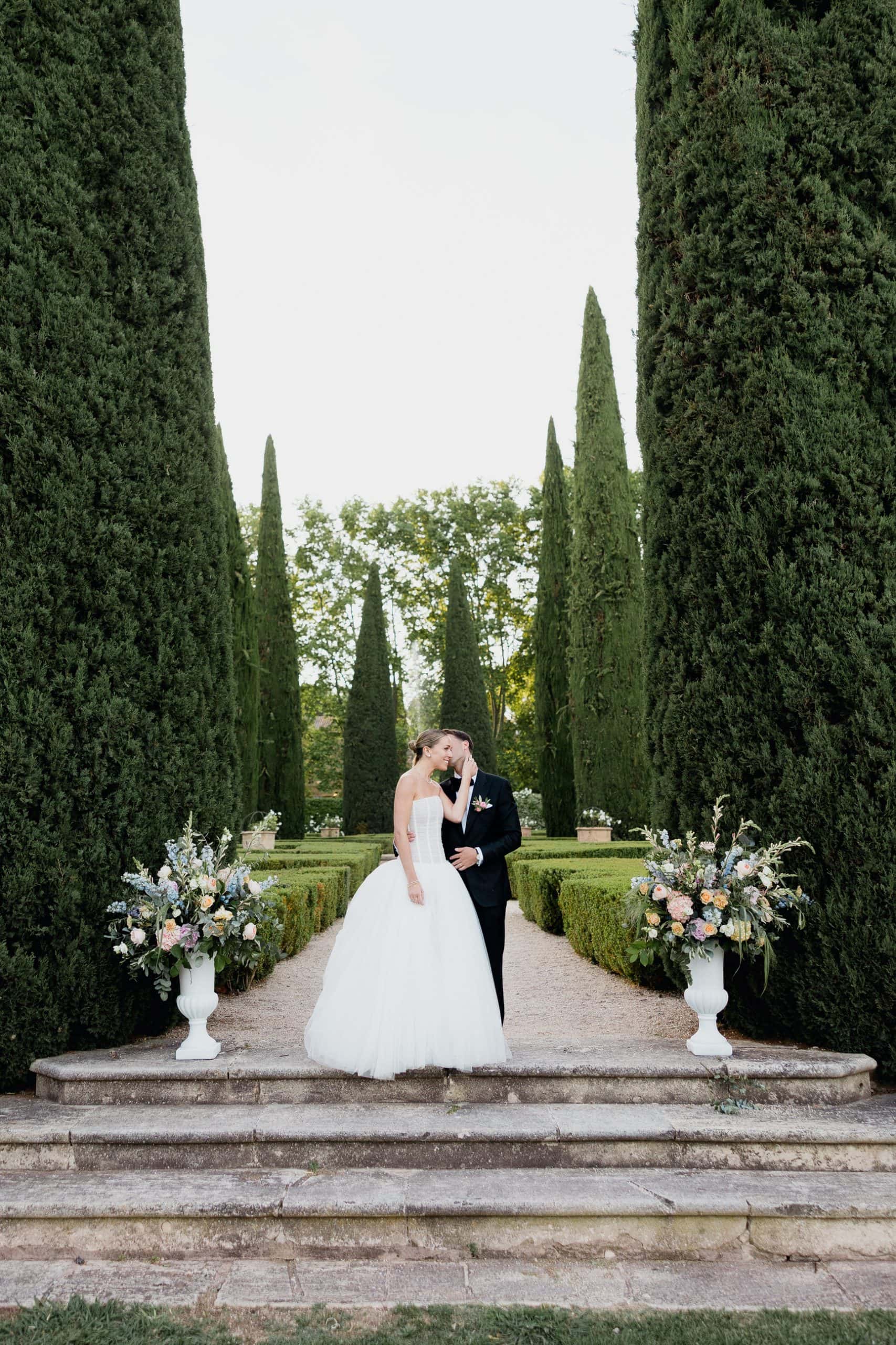 Cypress‑lined elegance at Château de Sannes – Maxime Bernadin. Bride in a voluminous gown and groom in a tuxedo stand among cypress trees at Château de Sannes, captured by Maxime Bernadin.