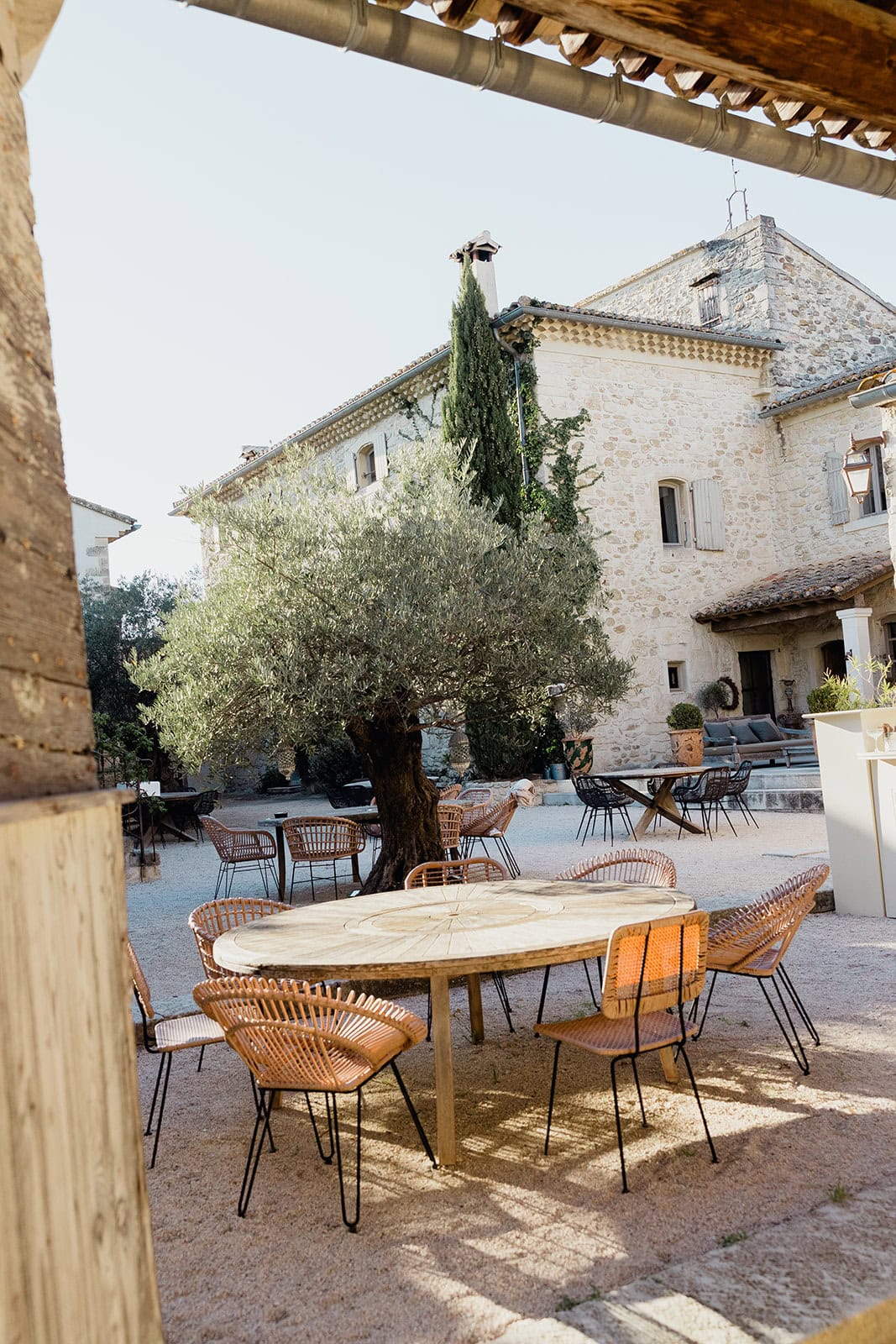 Tranquil courtyard at Domaine de Patras – Maxime Bernadin. Rustic courtyard with wooden tables and wicker chairs beneath an olive tree at Domaine de Patras, photographed by Maxime Bernadin.