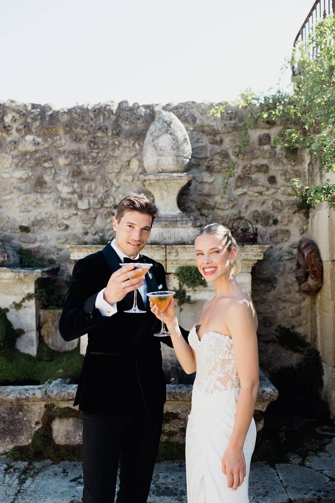 Celebratory toast at Domaine de Patras – Maxime Bernadin. Bride and groom toast with cocktails in a rustic stone courtyard at Domaine de Patras, captured by Maxime Bernadin.